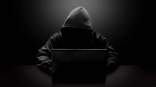 Ein Mann mit schwarzer Kapuze und verschattetem Gesicht sitzt vor einem Laptop.