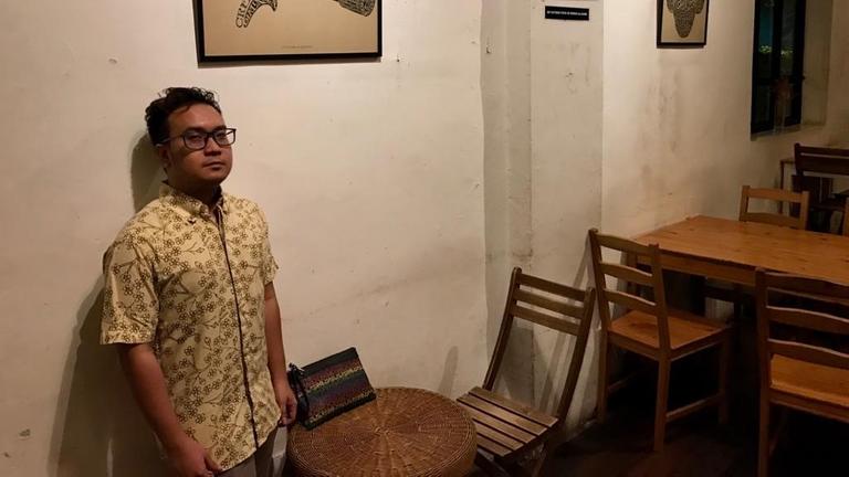 Ein junger Mann mit beigem Hemd und Brille lehnt an der Wand eines Cafés mit Holztischen und -stühlen.