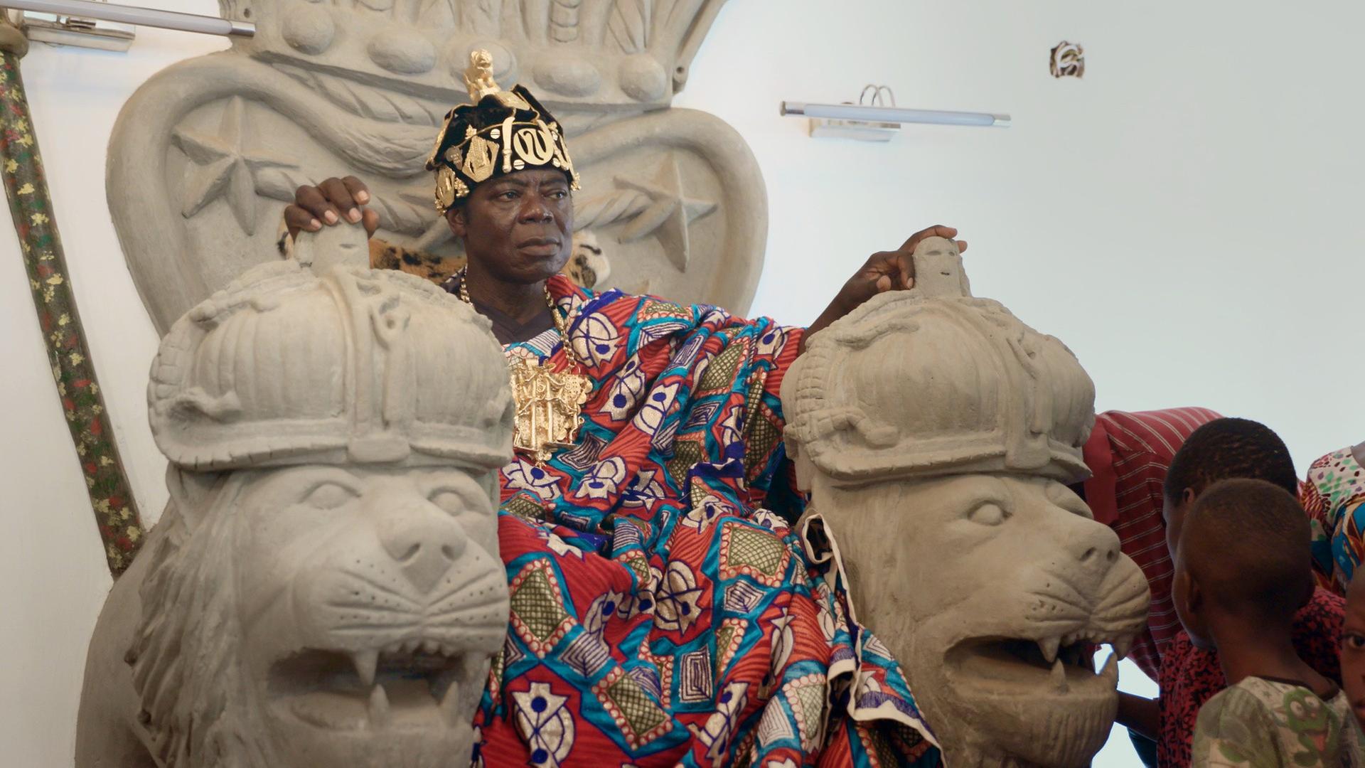 "König Bansah und seine Tochter": König Céphas Bansah sitzt in seiner traditionellen ghanaischen Königskleidung auf einem Thron aus behauenem Stein mit Löwenköpfen als Armlehmen.