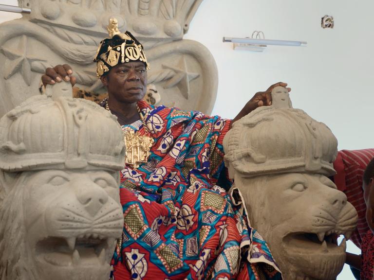 "König Bansah und seine Tochter": König Céphas Bansah sitzt in seiner traditionellen ghanaischen Königskleidung auf einem Thron aus behauenem Stein mit Löwenköpfen als Armlehmen.