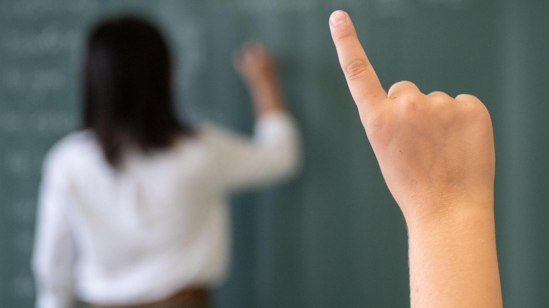 Hand eines Schülers einer fünften Klasse, der sich während einer Unterrichtsstunde meldet. Im Hintergrund des Bildes schreibt die Lehrerin an die Tafel.