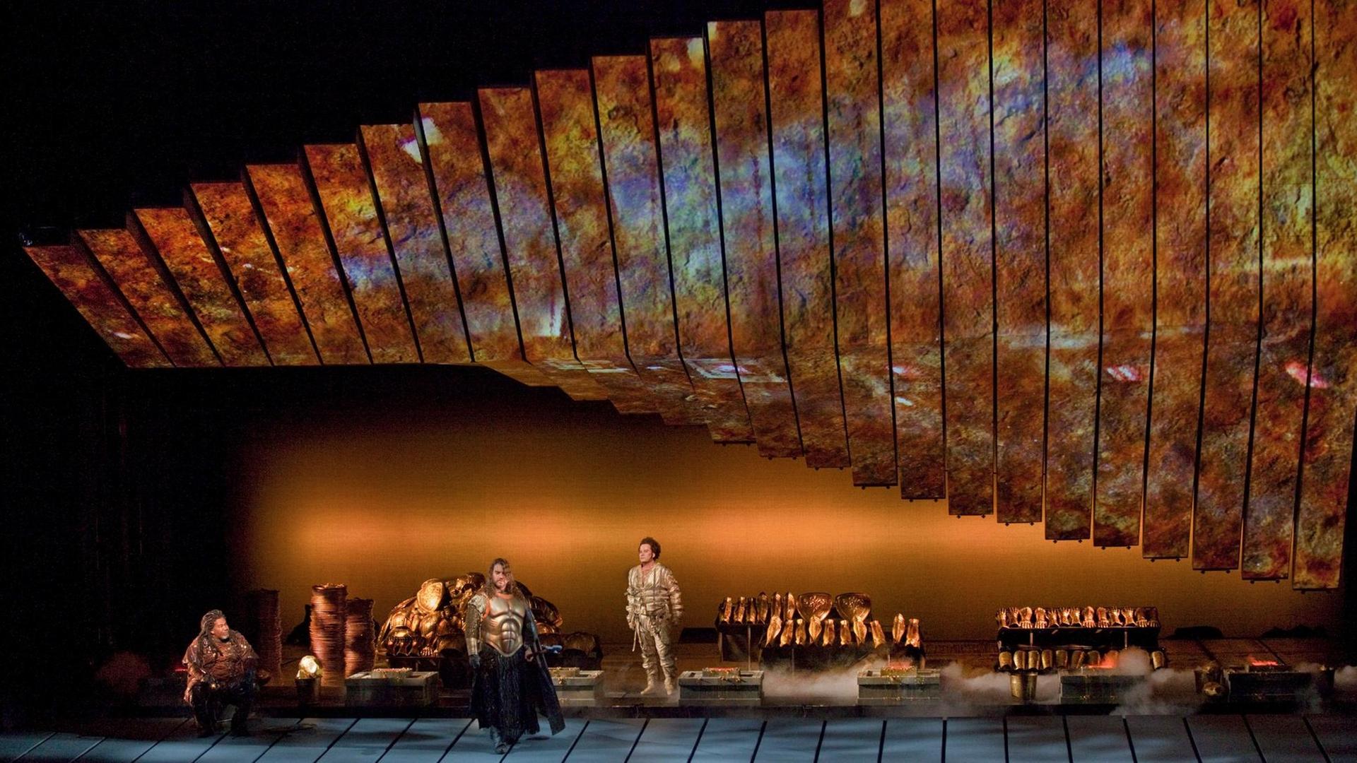 Eric Owens als Alberich, Bryn Terfel als Wotan, and Richard Croft als Loge in der Schmiede, in der viele goldene Teile aufbewahrt werden. Eric Owens als Alberich, Bryn Terfel als Wotan, and Richard Croft als Loge in der Schmiede, in der viele goldene Teile aufbewahrt werden.