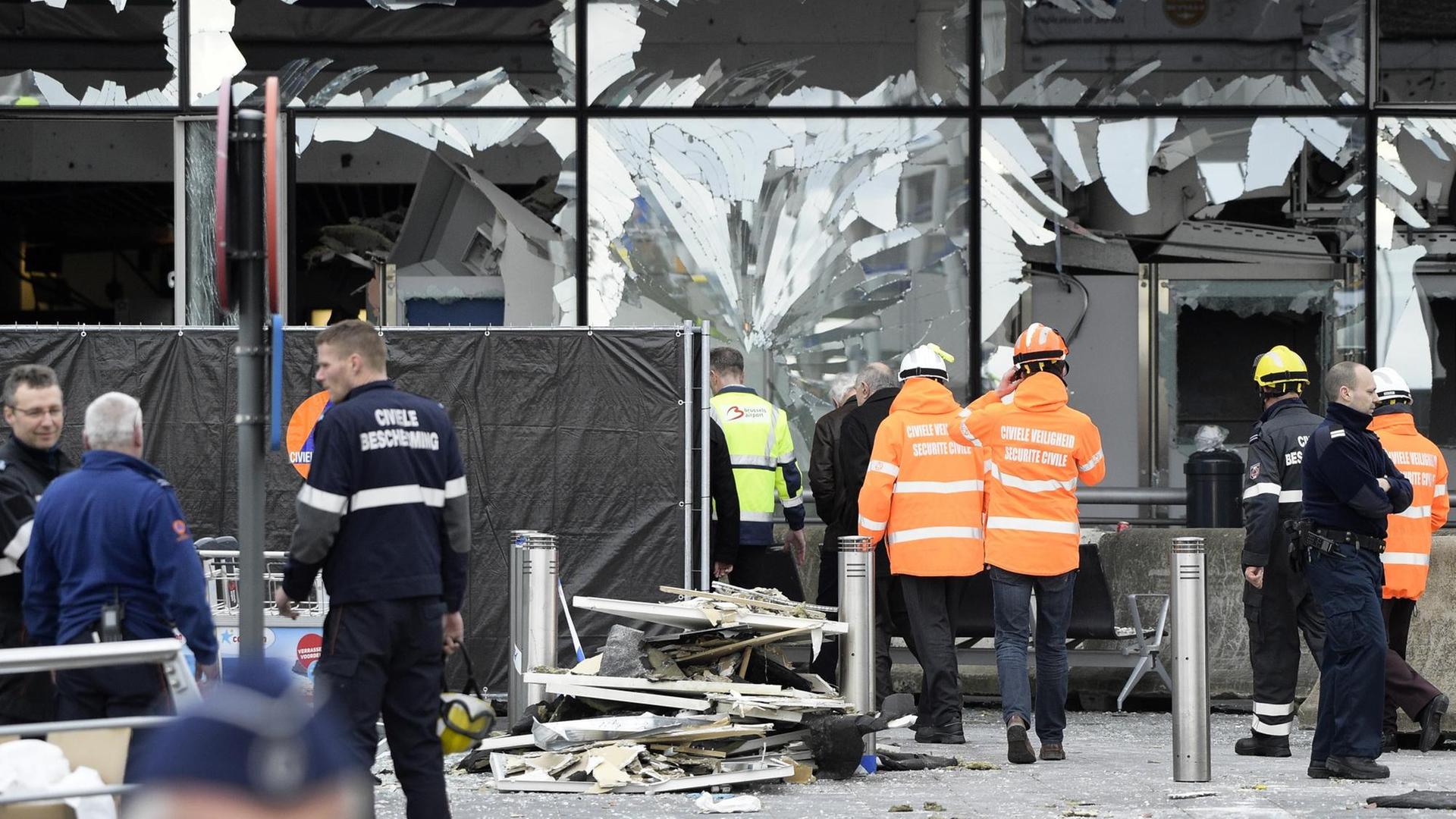 Zerborstene Scheiben am Terminal des Brüsseler Flughafens. Davor stehen Menschen mit Schutzhelmen und Warnwesten.