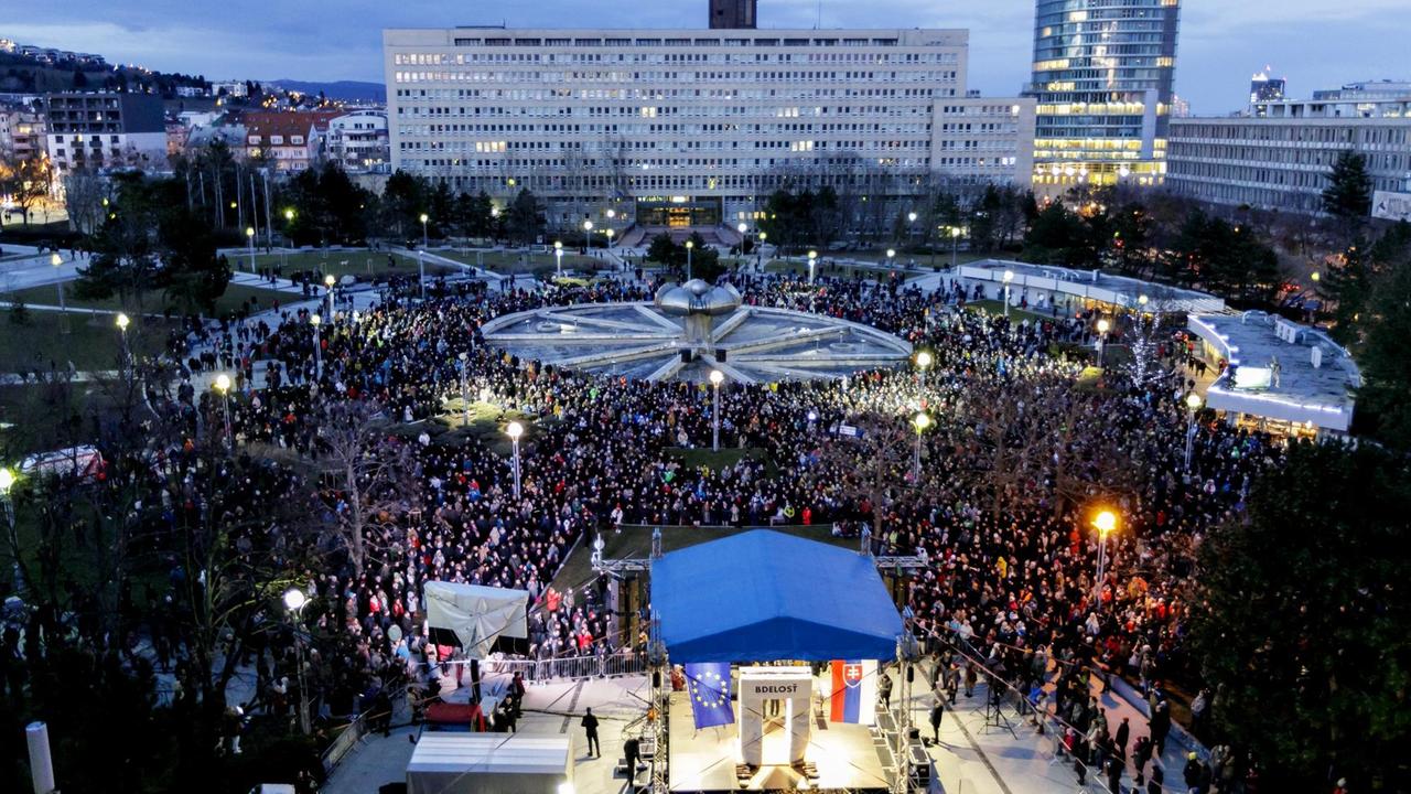 Blick aus größerer Höhe auf den Platz am Abend mit tausenden Demonstranten. Im Vordergrund eine beleuchtete Bühne, im Hintergrund Bürohäuser. 