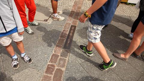 Junge Touristen stehen an Steinen, die auf dem Boden den Verlauf der ehemaligen Berliner Mauer markieren, an der Friedrichstraße nahe dem "Checkpoint Charlie" in Berlin im Bezirk Mitte.