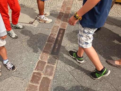 Junge Touristen stehen an Steinen, die auf dem Boden den Verlauf der ehemaligen Berliner Mauer markieren, an der Friedrichstraße nahe dem "Checkpoint Charlie" in Berlin im Bezirk Mitte.