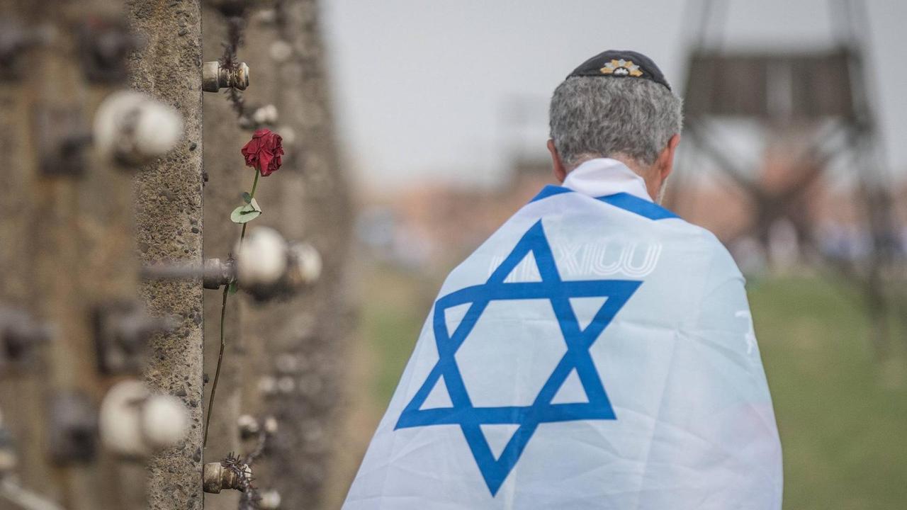 Ein in eine Israelfahne gehüllter Mann steht mit dem Rücken zur Kamera neben einer Wand im früheren Konzentrationslager Auschwitz, an der Gedenk-Devotionalien angebracht sind.