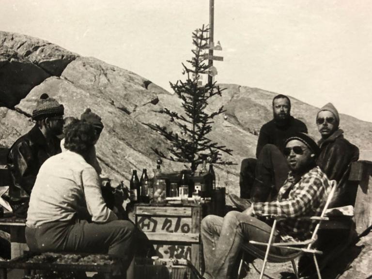 Alte Schwarzweiß-Aufnahme aus den 80er-Jahren, die zeigt, wie die Expeditionsteilnehmer in der Antarktis im Freien um eine als Tisch dienende Holzkiste herumsitzen. Darauf Flaschen und ein karger Weihnachtsbaum mit ein paar Christbaumkugeln.
