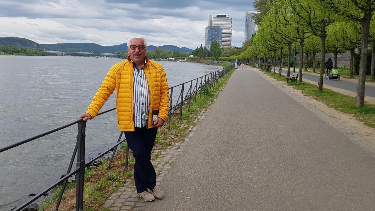 Heinz Hönig steht in gelber Jacke am Rheinufer. Im Hintergrund sind Hochhäuser des ehemaligen Regierungsviertels zu sehen.