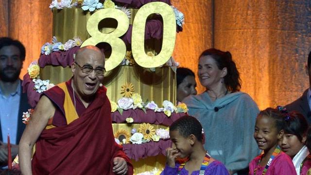 Der Dalai Lama vor einer riesigen Geburtstagstorte, umgeben von Kindern und Erwachsenen