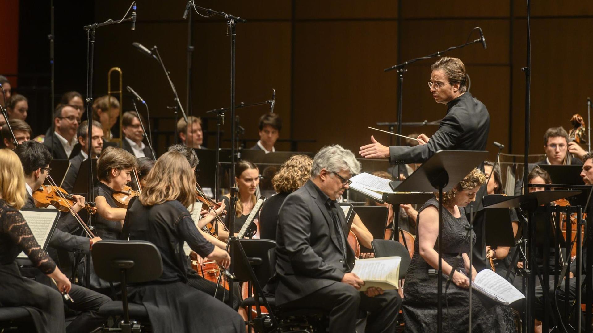 Ein Orchester mit Streichinstrumenten und ein Männerchor sitzen vor einem Dirigenten.