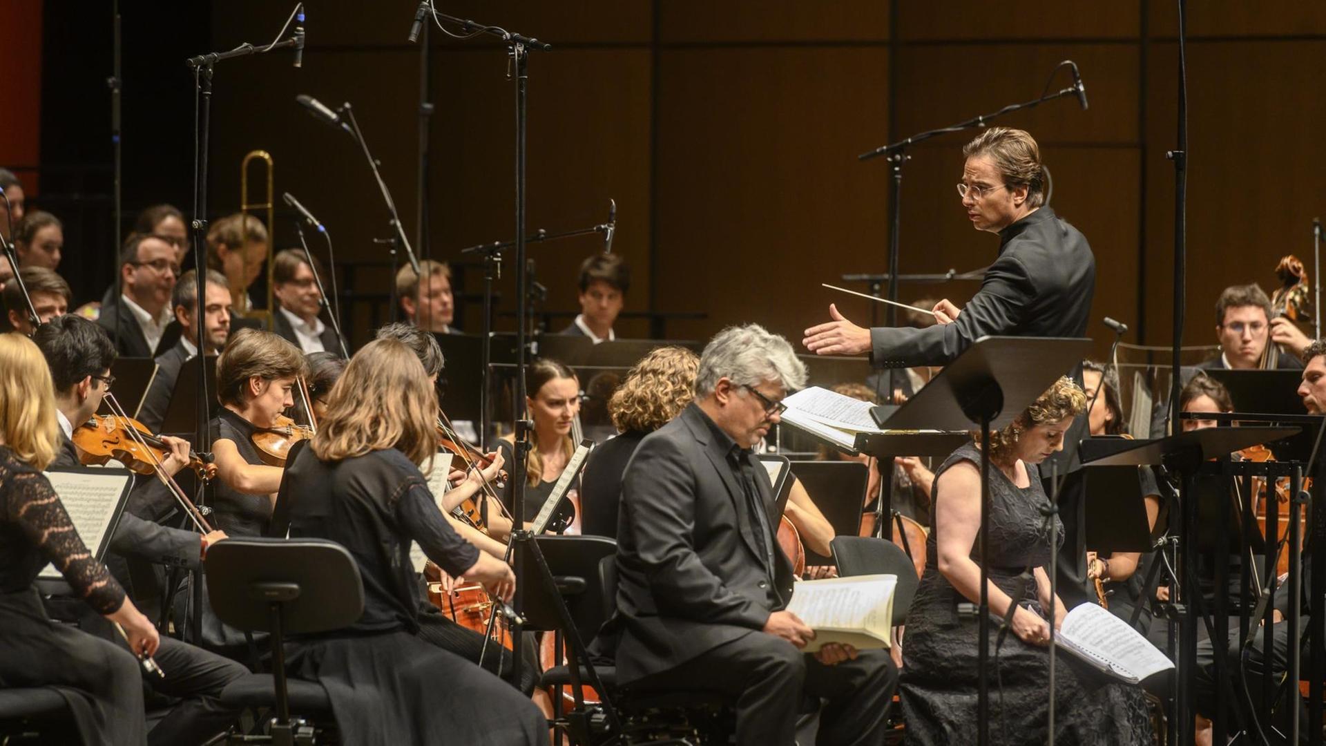 Ein Orchester mit Streichinstrumenten und ein Männerchor sitzen vor einem Dirigenten. Ein Orchester mit Streichinstrumenten und ein Männerchor sitzen vor einem Dirigenten.