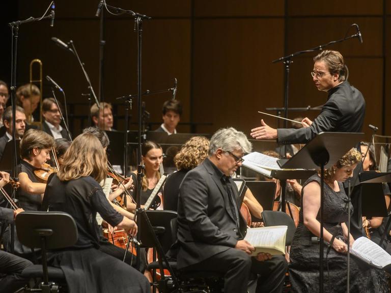 Ein Orchester mit Streichinstrumenten und ein Männerchor sitzen vor einem Dirigenten.