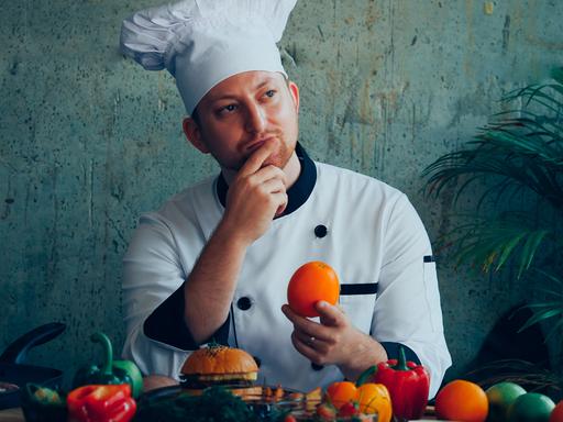 Ein junge Mann mit einer Kochmütze sitzt an einem Tisch, vor sich Obst und Gemüse. In der einen Hand hält er eine Orange, die andere Hand liegt an seinem Kinn.