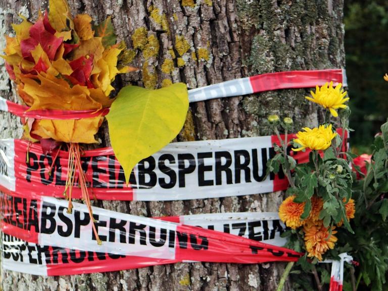 Blumen und Herbstlaub stecken zwischen den Absperrbändern der Polizei an der Dreisam in Freiburg, wo eine junge Frau getötet wurde.