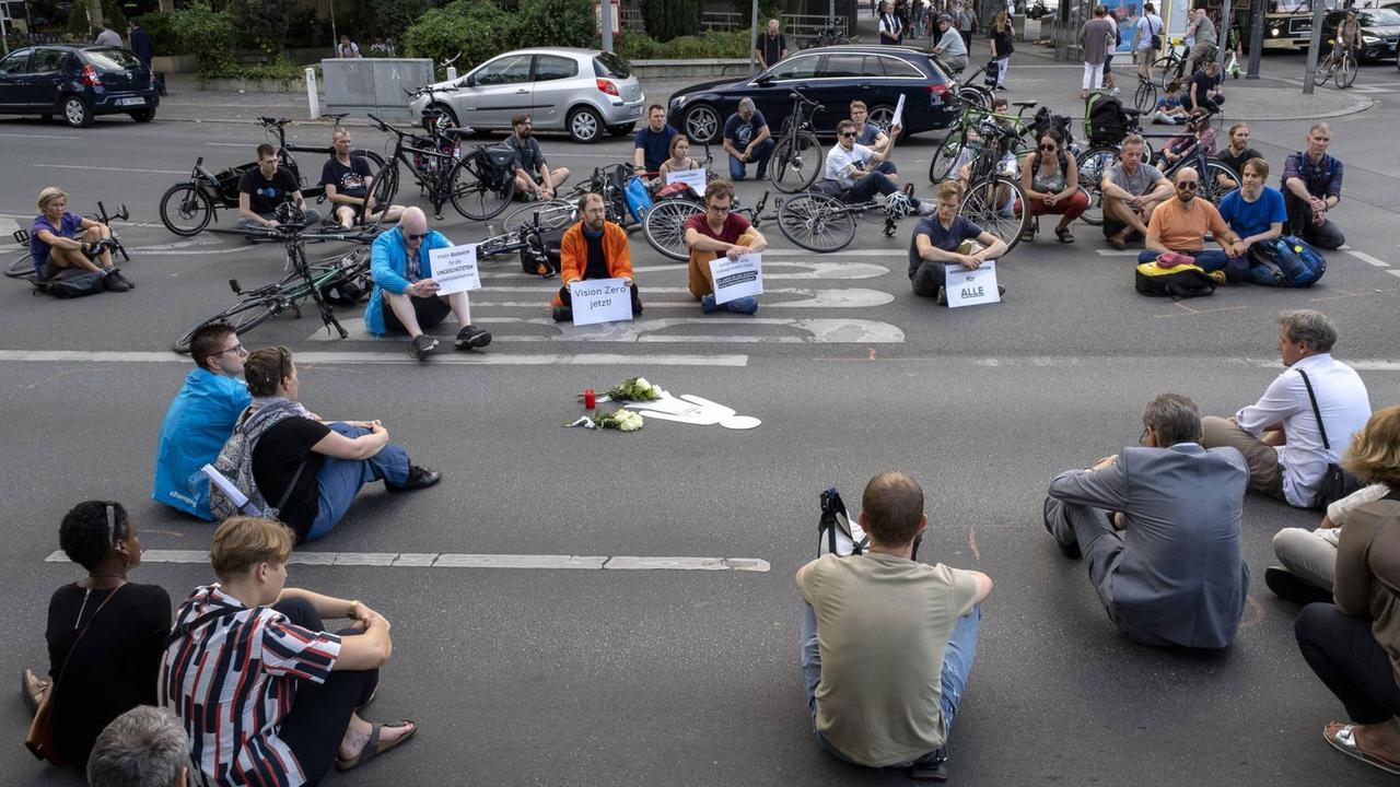 Mahnwache für eine getötete Fußgängerin in Berlin im Sommer 2018. Auf einer Kreuzung sitzen Menschen mit Blumen und Plakaten. Mahnwache für eine getötete Fußgängerin in Berlin im Sommer 2018. Auf einer Kreuzung sitzen Menschen mit Blumen und Plakaten.
