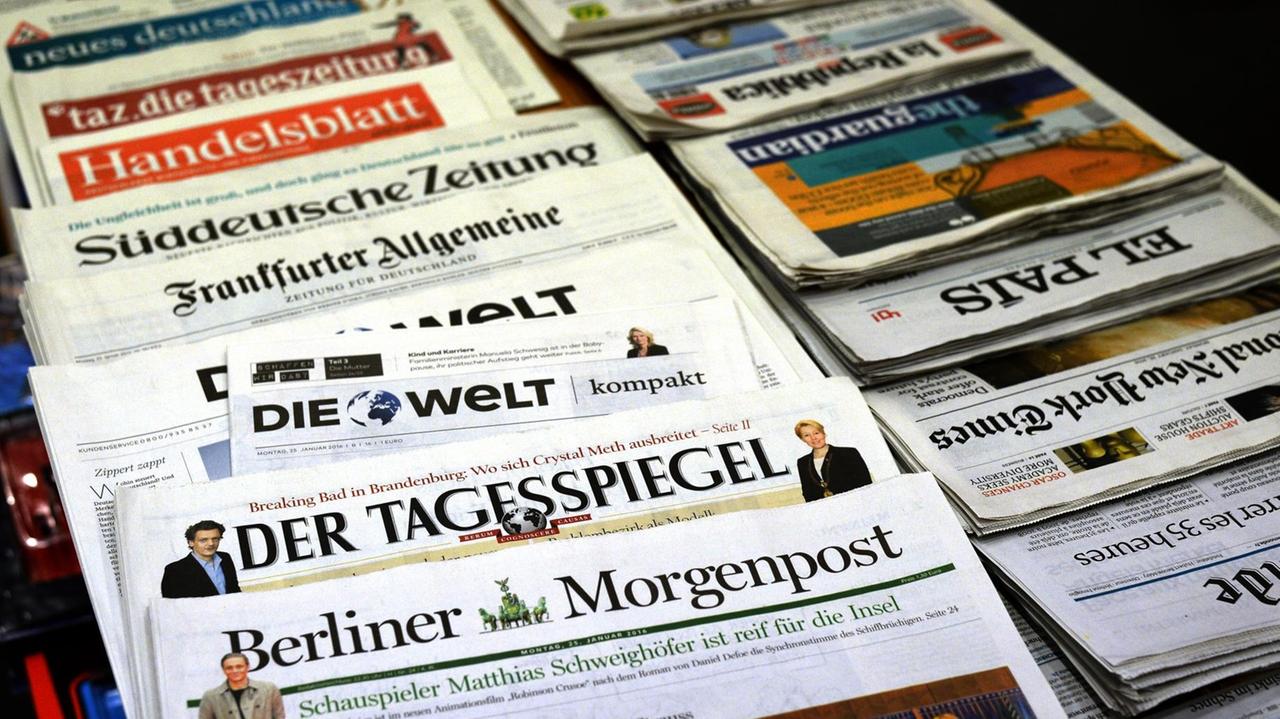In einer Reihe liegen deutsche Zeitungen (vorne die Berliner Morgenpost, der Tagesspiegel und die Welt). Daneben kopfüber eine Reihe ausländischer Blätter. 