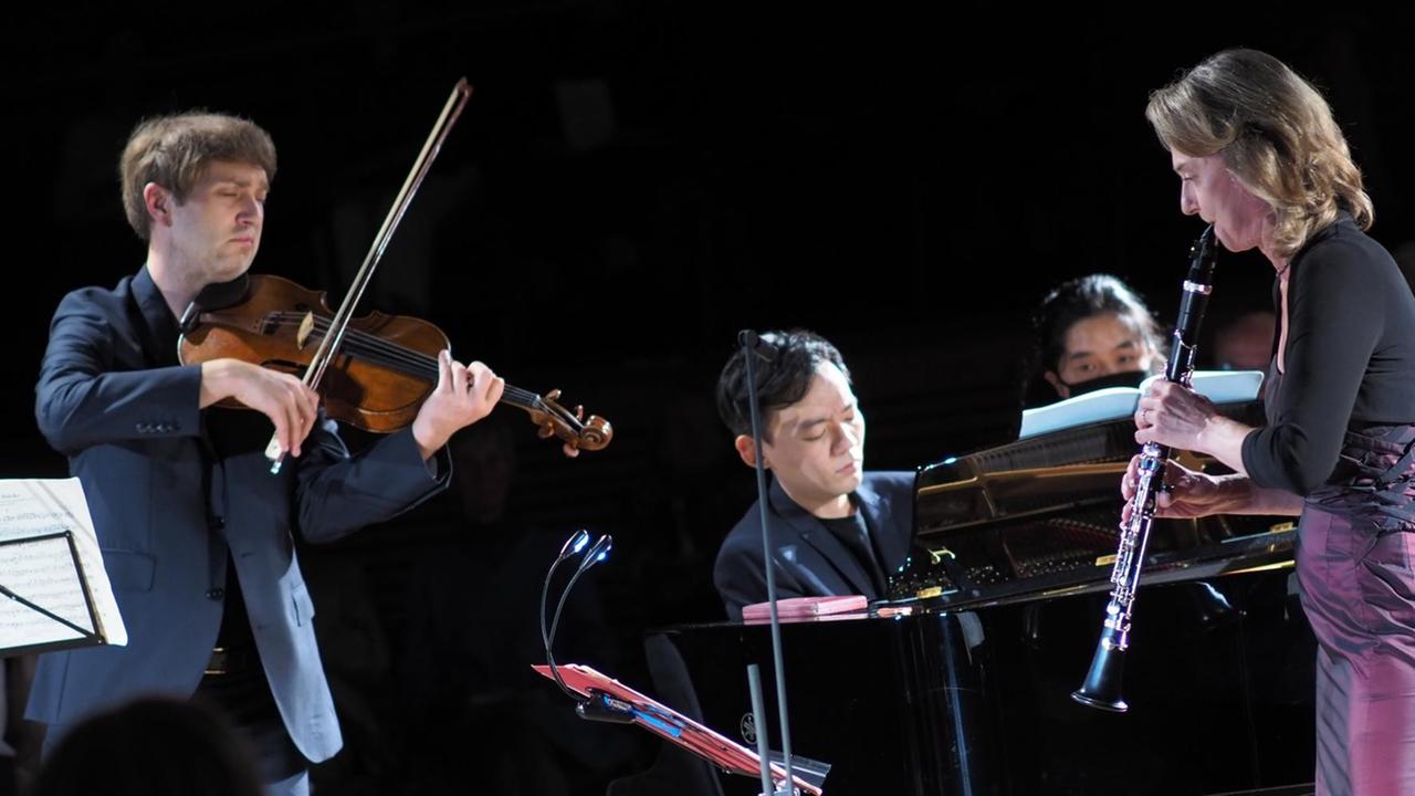 Drei Musiker, Bratsche, Klavier und Klarinette spielend, musizieren konzentriert zwischen Notenständern und Mikrofonen.