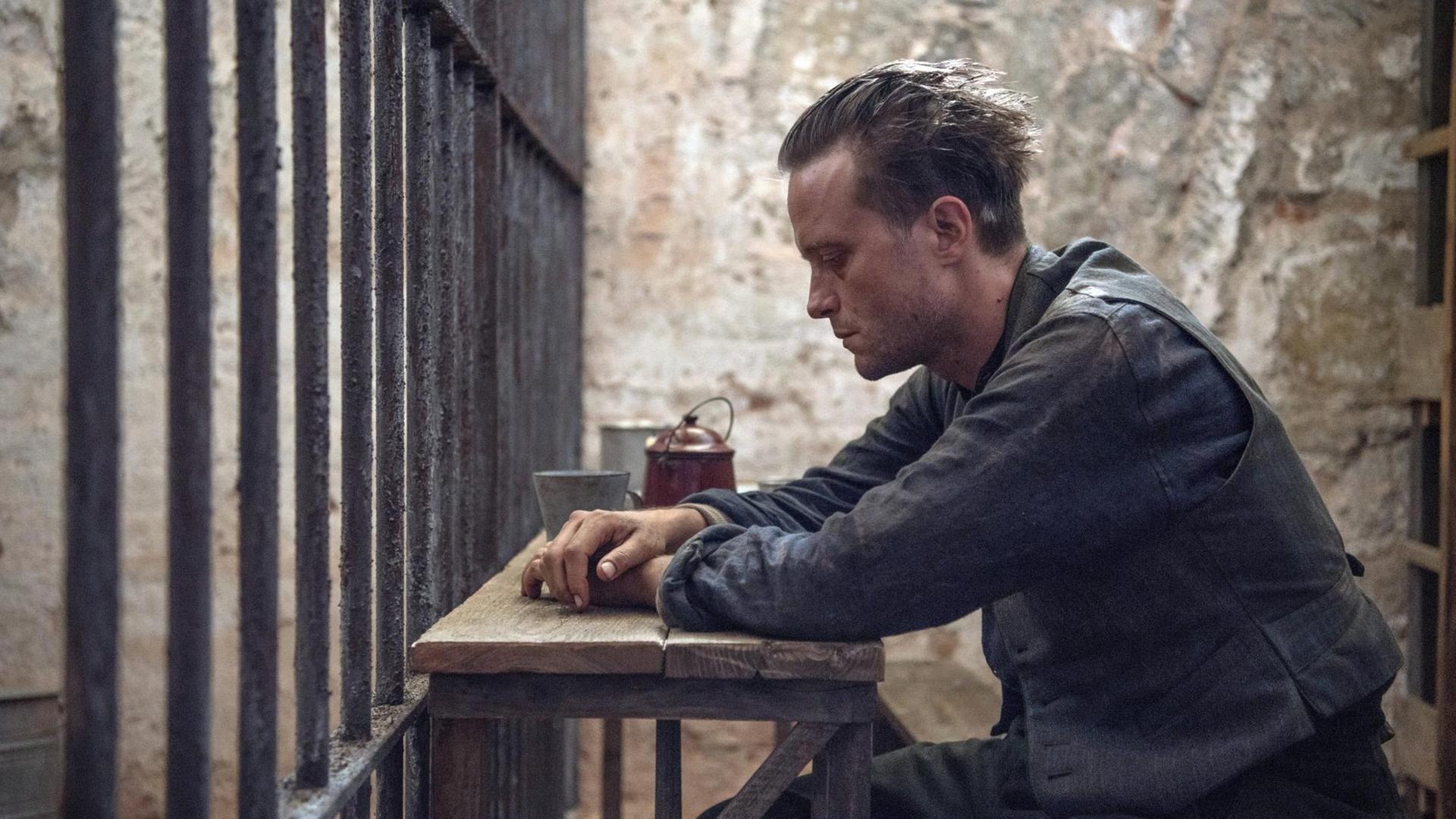 ein Mann sitzt an einem kargen Holztisch hinter Gittern