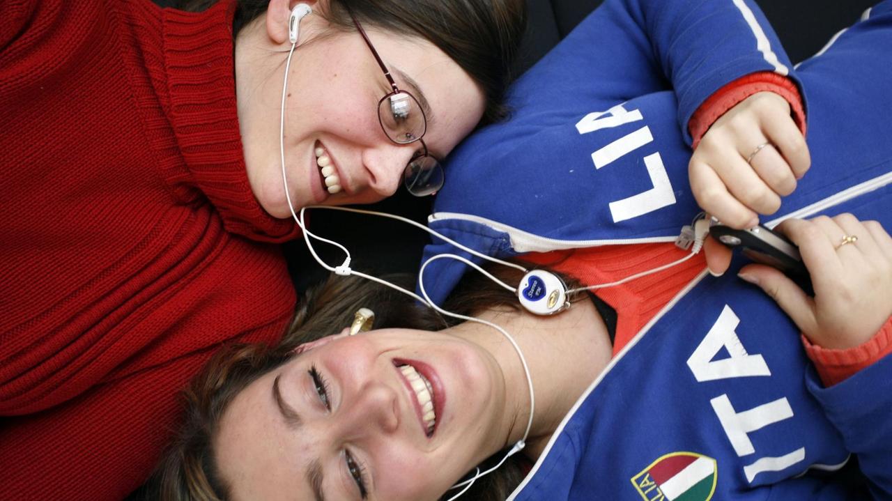 Zwei Frauen liegen Kopf an Kopf und teilen sich Ohrstöpsel. Eine trägt einen roten Pullover und Brille, die andere eine Trainingsjacke in blau mit der Aufschrift ITALIA. Zwei Frauen liegen Kopf an Kopf und teilen sich Ohrstöpsel. Eine trägt einen roten Pullover und Brille, die andere eine Trainingsjacke in blau mit der Aufschrift ITALIA.