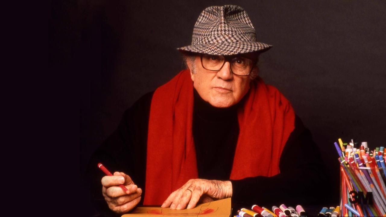 Der Regisseur Frederico Fellini mit Hut und rotem Schal skizziert etwas auf einem gelben mit Buntstiften.