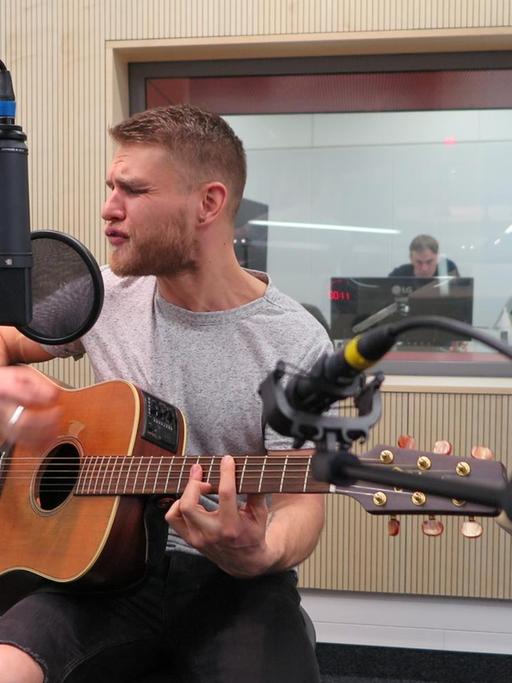 Ein Mann sitzt in einem Hörfunkstudio vor Mikrofonen, er spielt akustische Gitarre und singt mit geschlossenen Augen.