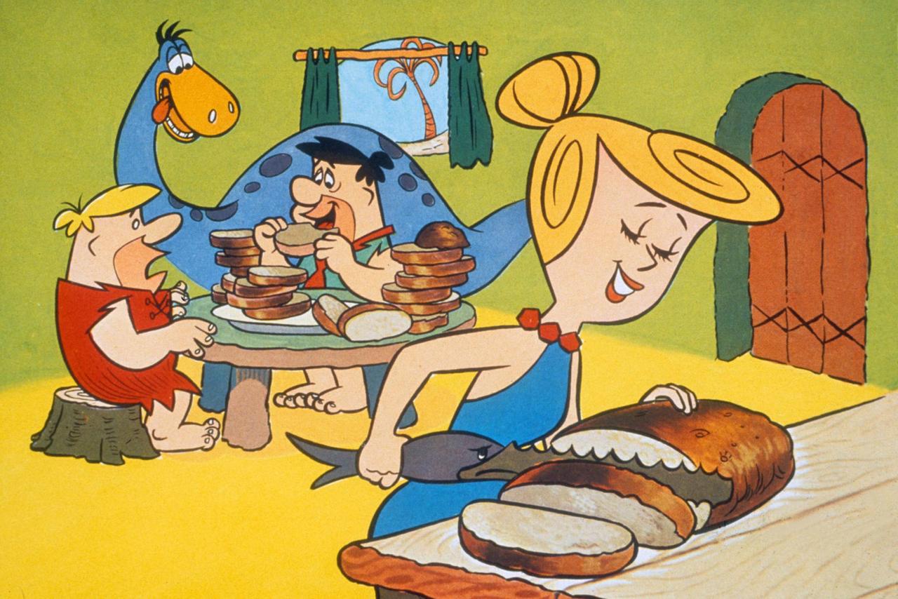 Szene aus der TV-Zeichentrickserie "Flintstones": Barney Rubble und Fred Flintstone unterhalten sich, Wilma Flintstone schneidet Brot mit einem Haifisch-Messer. Szene aus der TV-Zeichentrickserie "Flintstones": Barney Rubble und Fred Flintstone unterhalten sich, Wilma Flintstone schneidet Brot mit einem Haifisch-Messer.