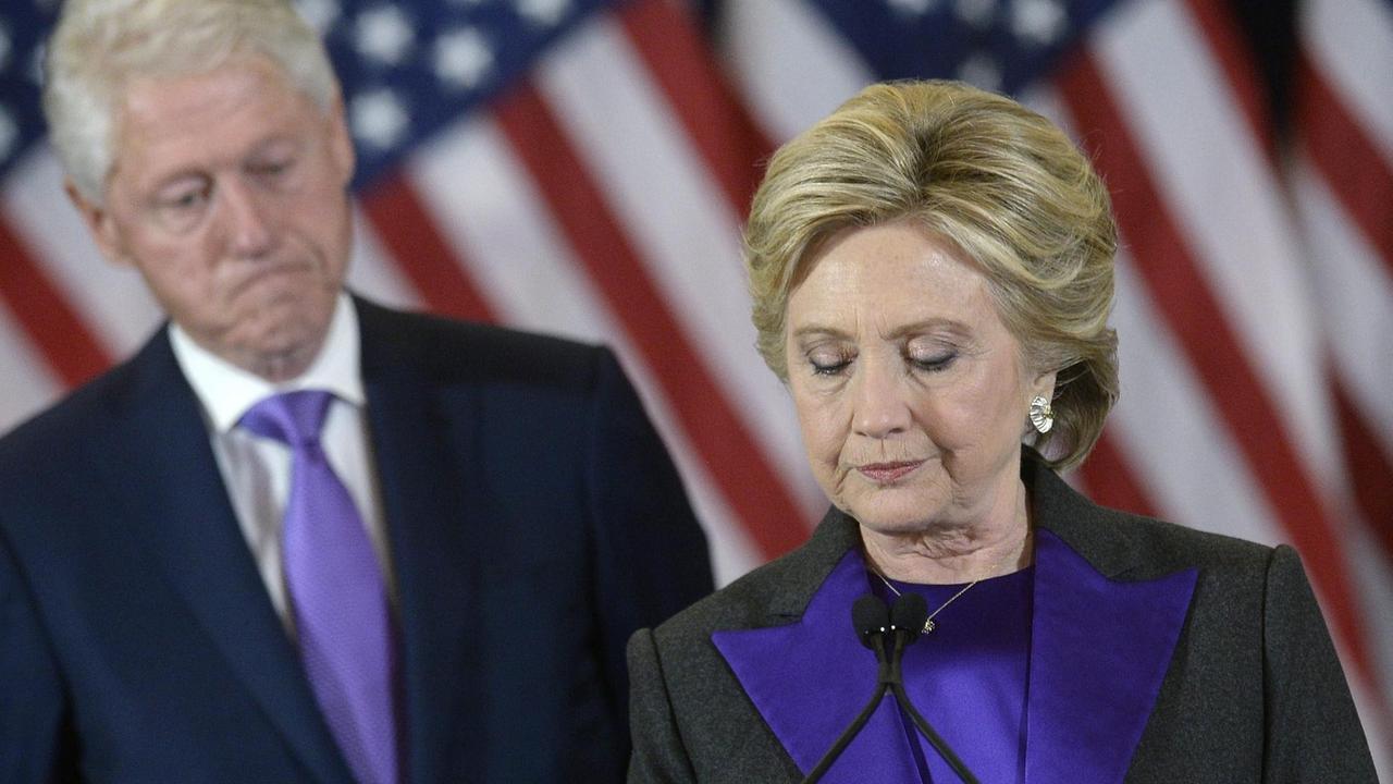 Hillary Clinton steht vor US-Fahnen. Im Hintergrund ist ihr Mann Bill zu sehen.