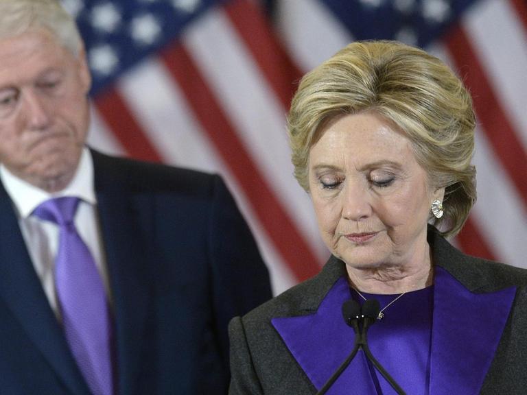 Hillary Clinton steht vor US-Fahnen. Im Hintergrund ist ihr Mann Bill zu sehen.