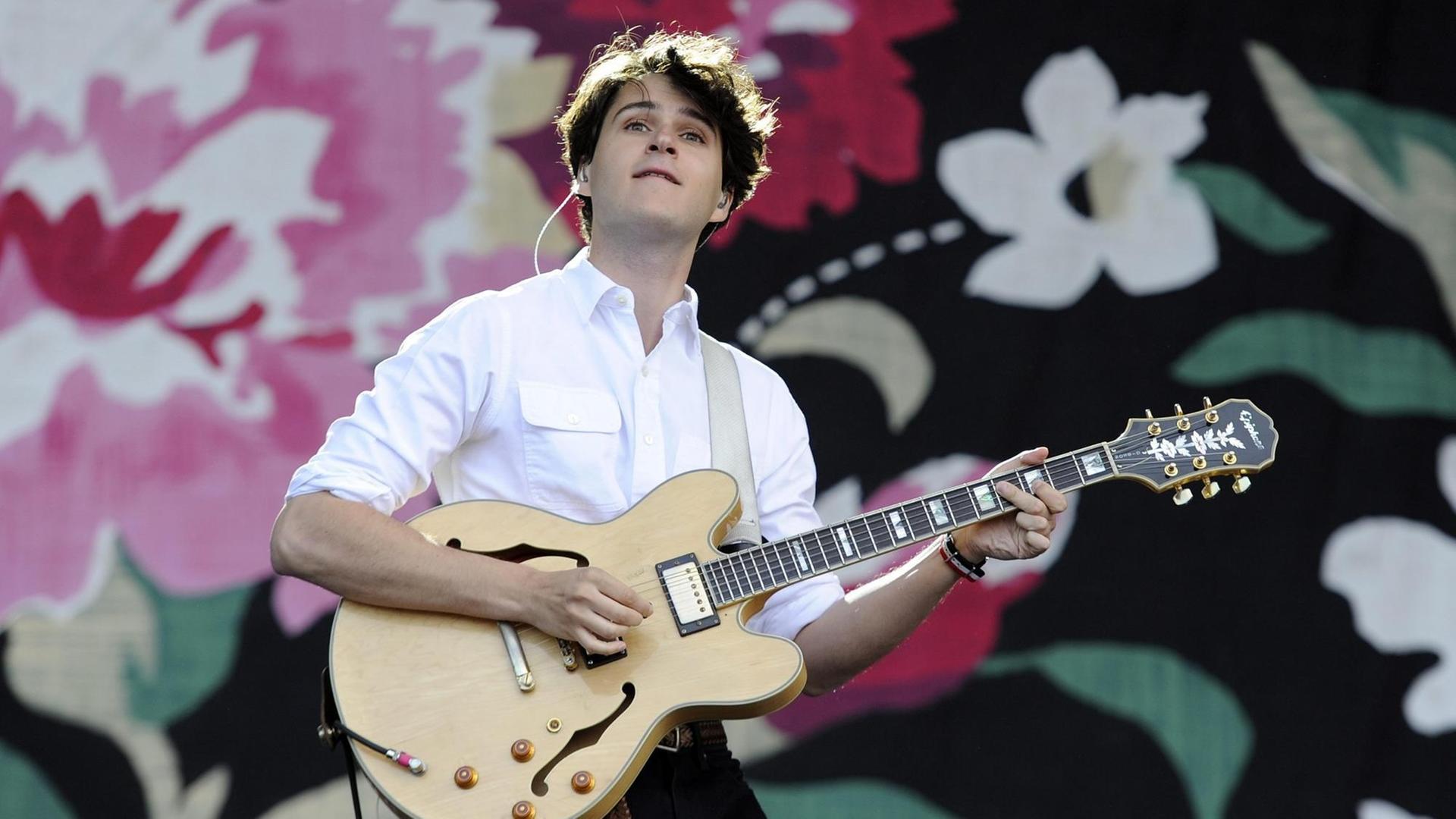 Der Frontmann von Vampire Weekend, Ezra Koenig, in weißem Hemd und mit Gitarre vor einem geblümten Hintergrund beim Glastonbury Festival 2013 Der Frontmann von Vampire Weekend, Ezra Koenig, in weißem Hemd und mit Gitarre vor einem geblümten Hintergrund beim Glastonbury Festival 2013
