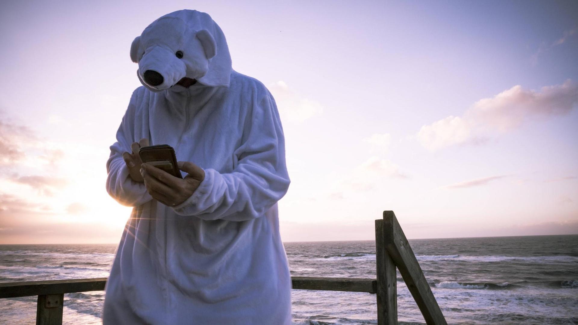 Ein Mann steht am Strand, trägt ein Eisbärkostüm und hält Smartphone in der Hand. Ein Mann steht am Strand, trägt ein Eisbärkostüm und hält Smartphone in der Hand.
