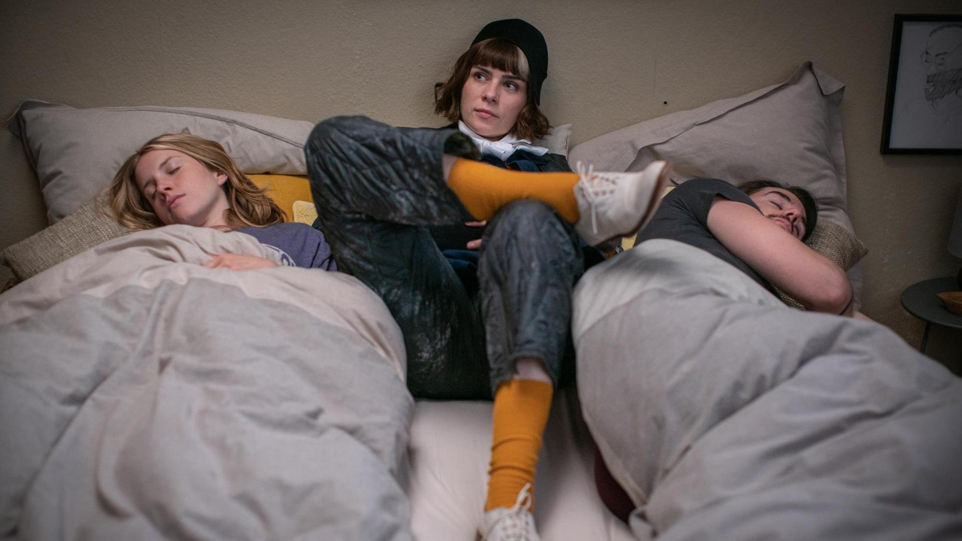 Filmszene aus "The Mopes", Monika (Nora Tschirner) liegt voll bekleidet zwischen dem schlagenden Mat (Roel Dirven) und der schlafenden Susa (Paula Kalenberg) im Bett. Filmszene aus "The Mopes", Monika (Nora Tschirner) liegt voll bekleidet zwischen dem schlagenden Mat (Roel Dirven) und der schlafenden Susa (Paula Kalenberg) im Bett.