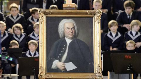 Ein golden gerahmtes Gemälde von Bach steht vor dem aufgestellten Thomanerchor. Das Blatt, was er in der Hand hält, ist leer, ganz ohne Noten.
