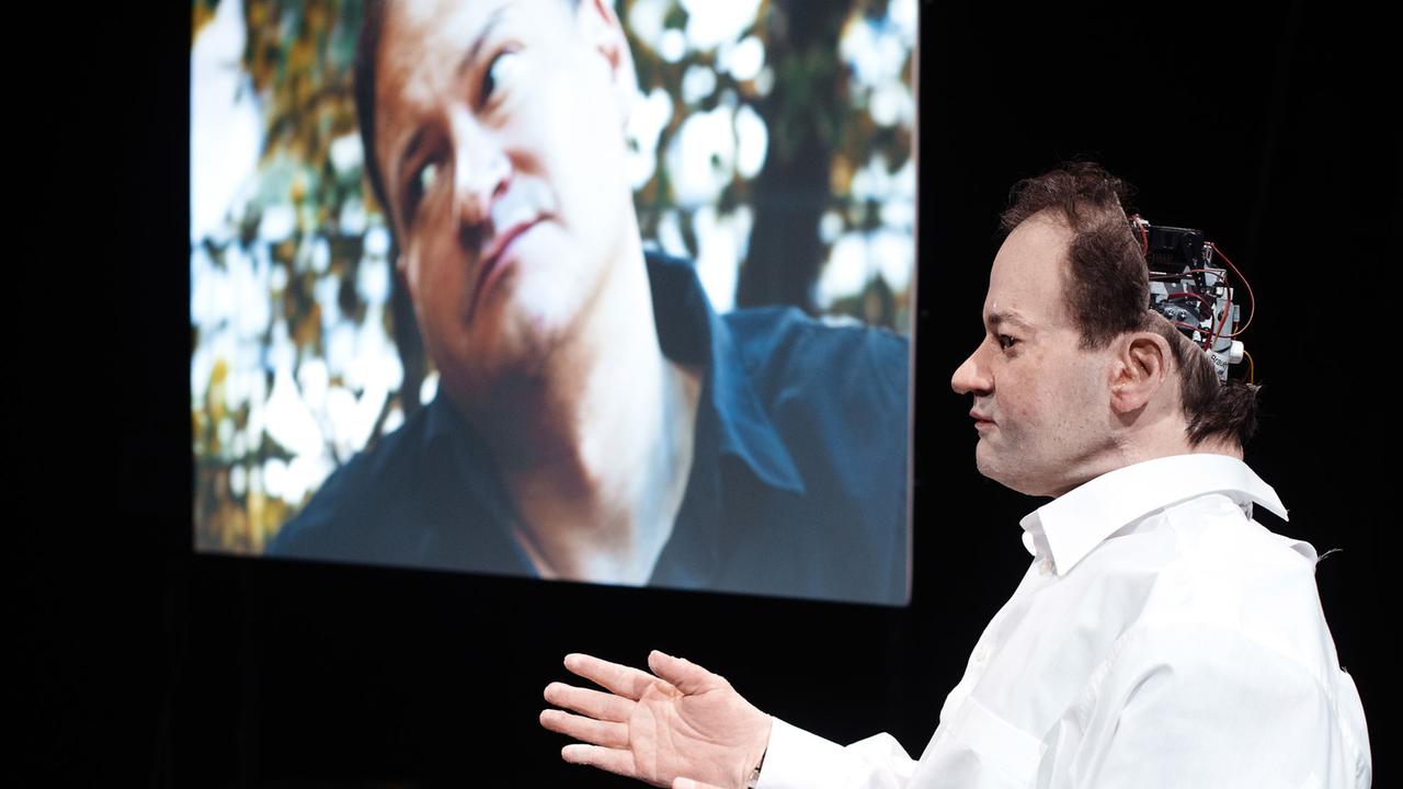 Der Autor Thomas Melle sitzt als Roboter auf einem Sessel, im Hintergrund ist der echte Melle in einem Video zu sehen. Foto: Gabriela Neeb Der Autor Thomas Melle sitzt als Roboter auf einem Sessel, im Hintergrund ist der echte Melle in einem Video zu sehen. Foto: Gabriela Neeb