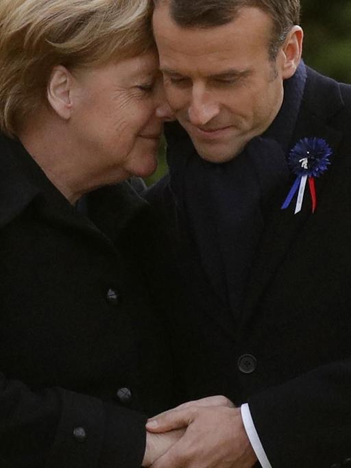 Merkel legt ihren Kopf mit geschlossenen Augen an den Kopf Macrons.
