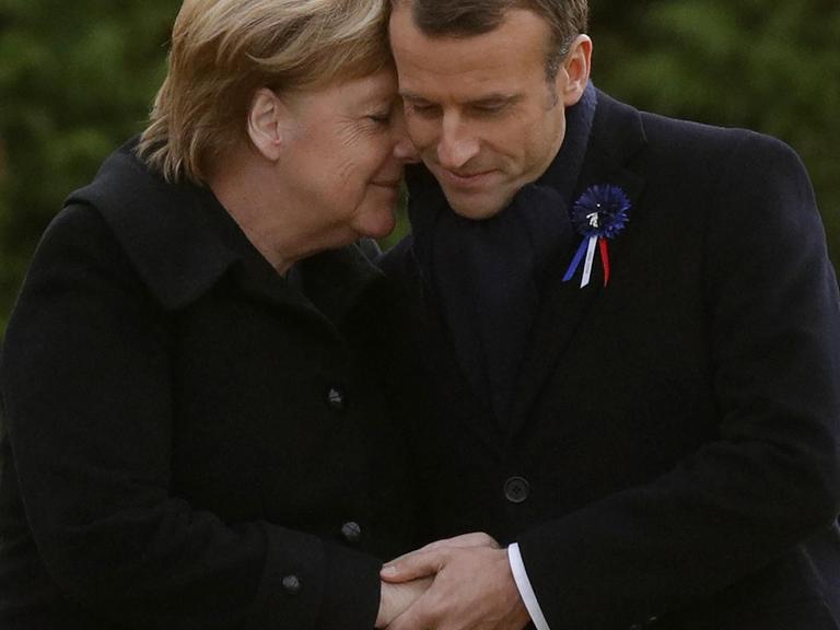 Merkel legt ihren Kopf mit geschlossenen Augen an den Kopf Macrons.