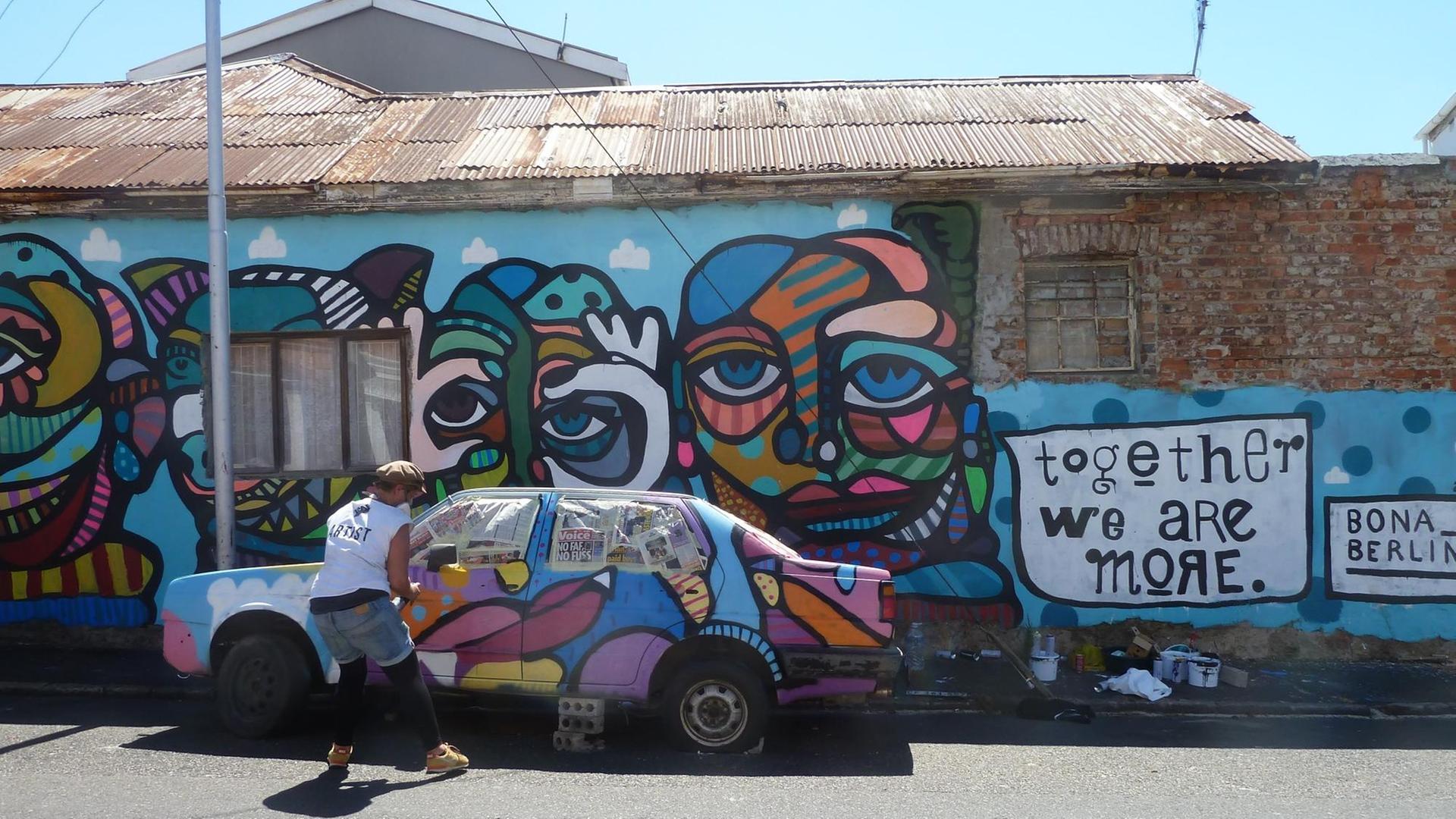 Ein buntes Graffiti aus Gesichtern an einer Häuserwand und über einem geparkten Auto. Rechts ebenfalls aufgesprayt der Spruch "Together We Are More". Ein buntes Graffiti aus Gesichtern an einer Häuserwand und über einem geparkten Auto. Rechts ebenfalls aufgesprayt der Spruch "Together We Are More".