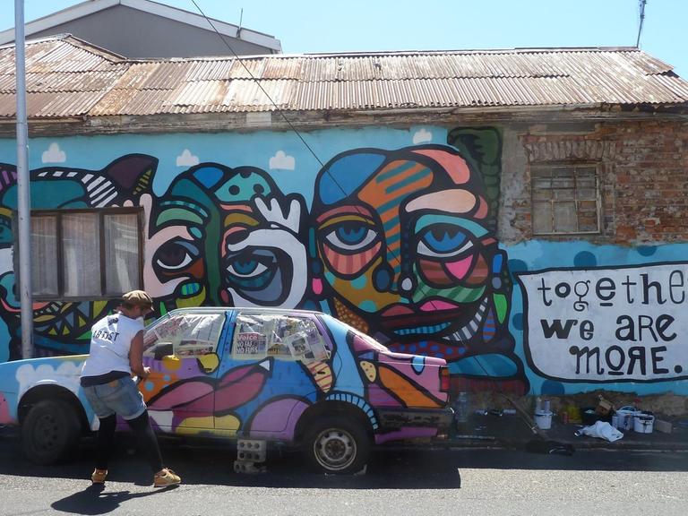 Ein buntes Graffiti aus Gesichtern an einer Häuserwand und über einem geparkten Auto. Rechts ebenfalls aufgesprayt der Spruch "Together We Are More".