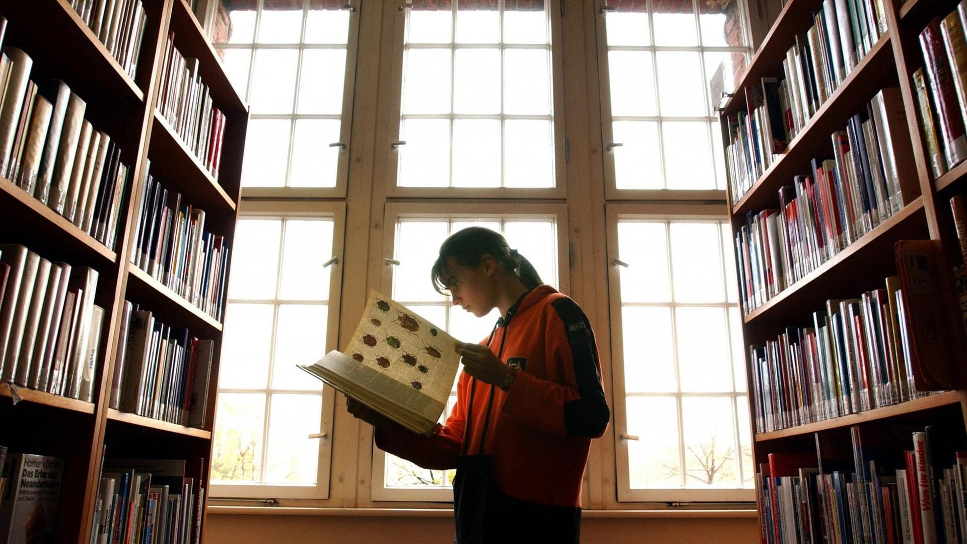 Eine Frau steht in einer Bibliothek zwischen zwei Regalen und schaut in ein aufgeschlagenes Buch