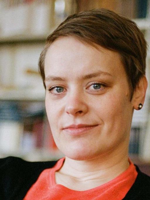 Hanna Engelmeier sitzt vor einer Bücherwand. Sie trägt ein hellrotes Oberteil und eine schwarze Jacke und schaut in die Kamera. Das Haar trägt sie kurz mit Seitenscheitel.