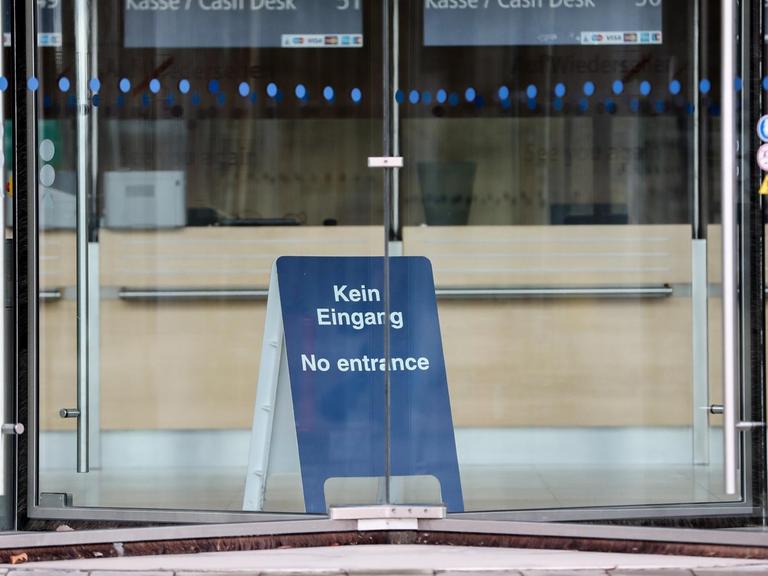 Durch eine Drehtür ist ein blaues Aufstellschild mit der Aufschrift 'Kein Eingang, No entrance' zu sehen. Im Hintergrund sind die leeren Kassenplätze.