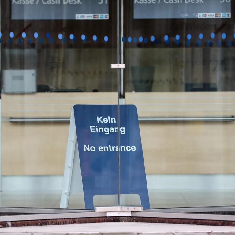 Durch eine Drehtür ist ein blaues Aufstellschild mit der Aufschrift 'Kein Eingang, No entrance' zu sehen.
Im Hintergrund sind die leeren Kassenplätze.