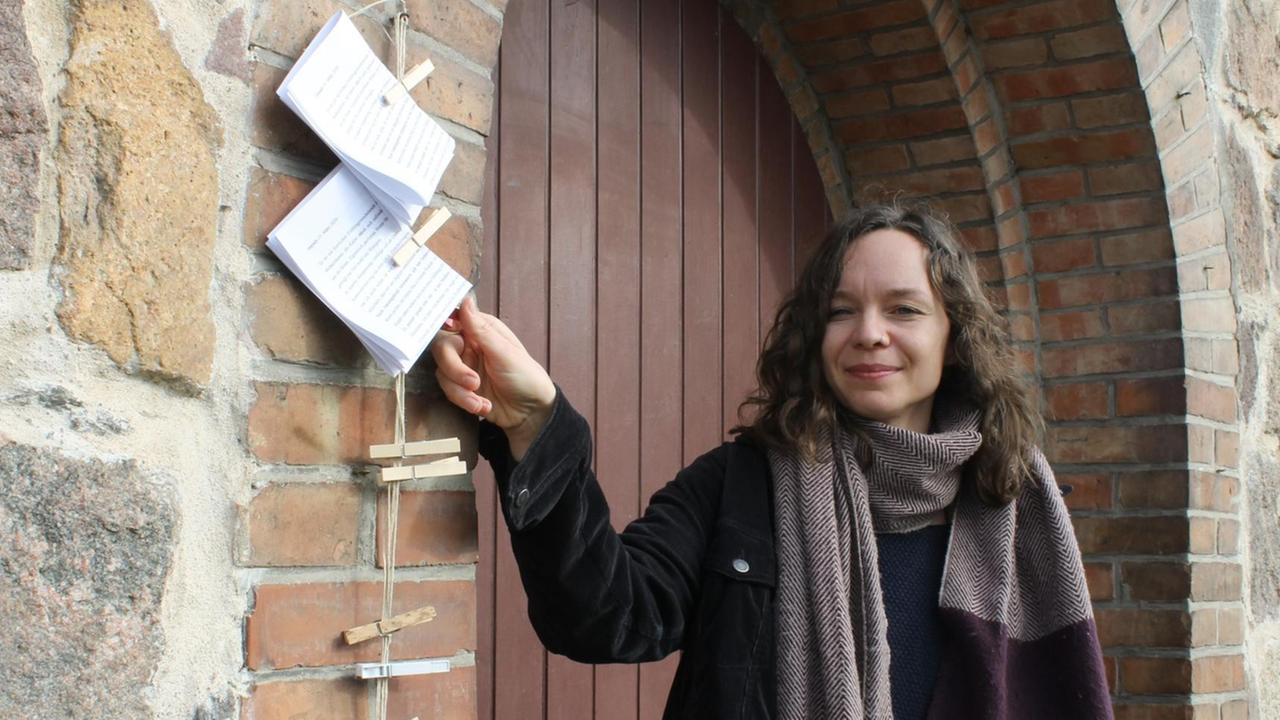 Pfarrerin Susanne Brusch, steht vor der Kirchentür und zeigt die an einer Wäscheleine aufgehängten Zettel mit Texten, die Gemeindemitgliedern in der Corona-Krise Mut machen sollen.