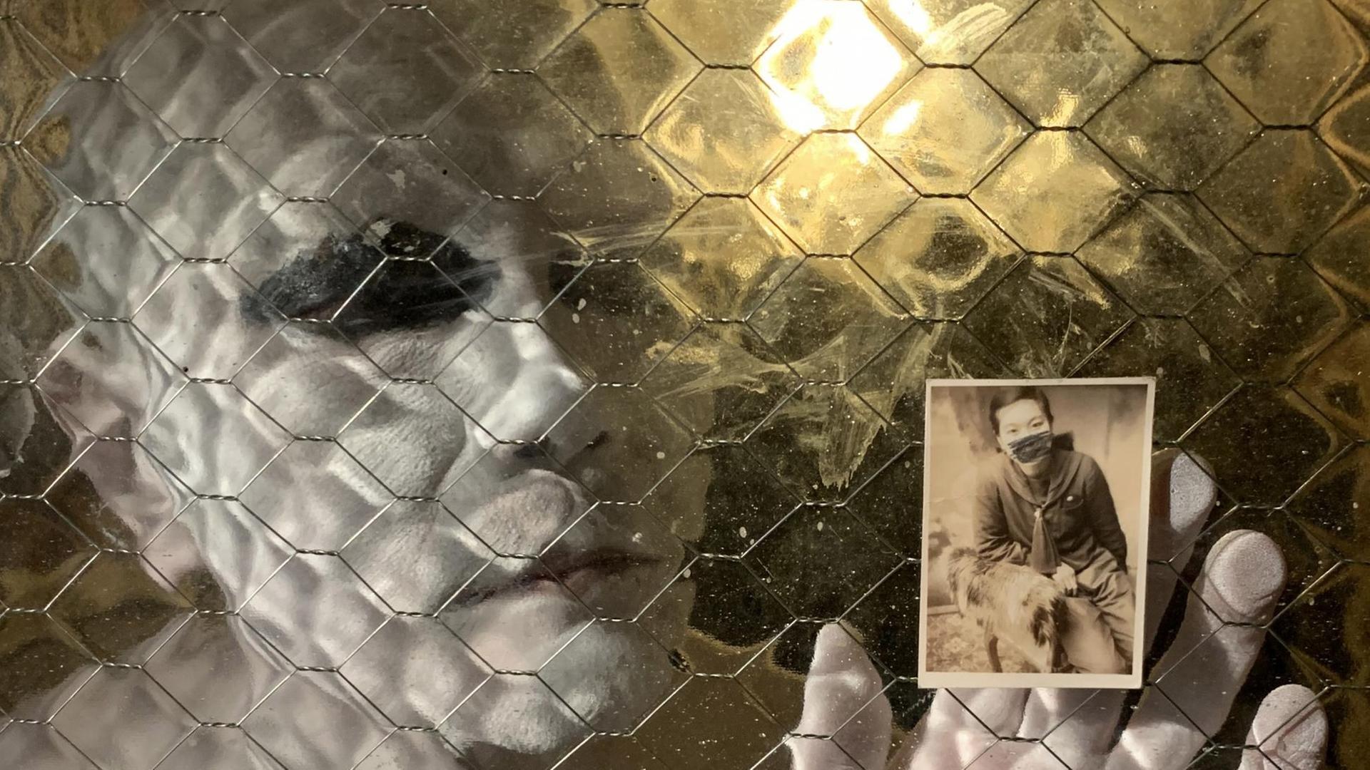Durchblick durch ein Fenster auf einen mit weißer Hautfarbe bemalten Menschen ohne Kopfhaar, der ein Schwarzweißfoto einer Person gegen die Scheibe hält. Durchblick durch ein Fenster auf einen mit weißer Hautfarbe bemalten Menschen ohne Kopfhaar, der ein Schwarzweißfoto einer Person gegen die Scheibe hält.