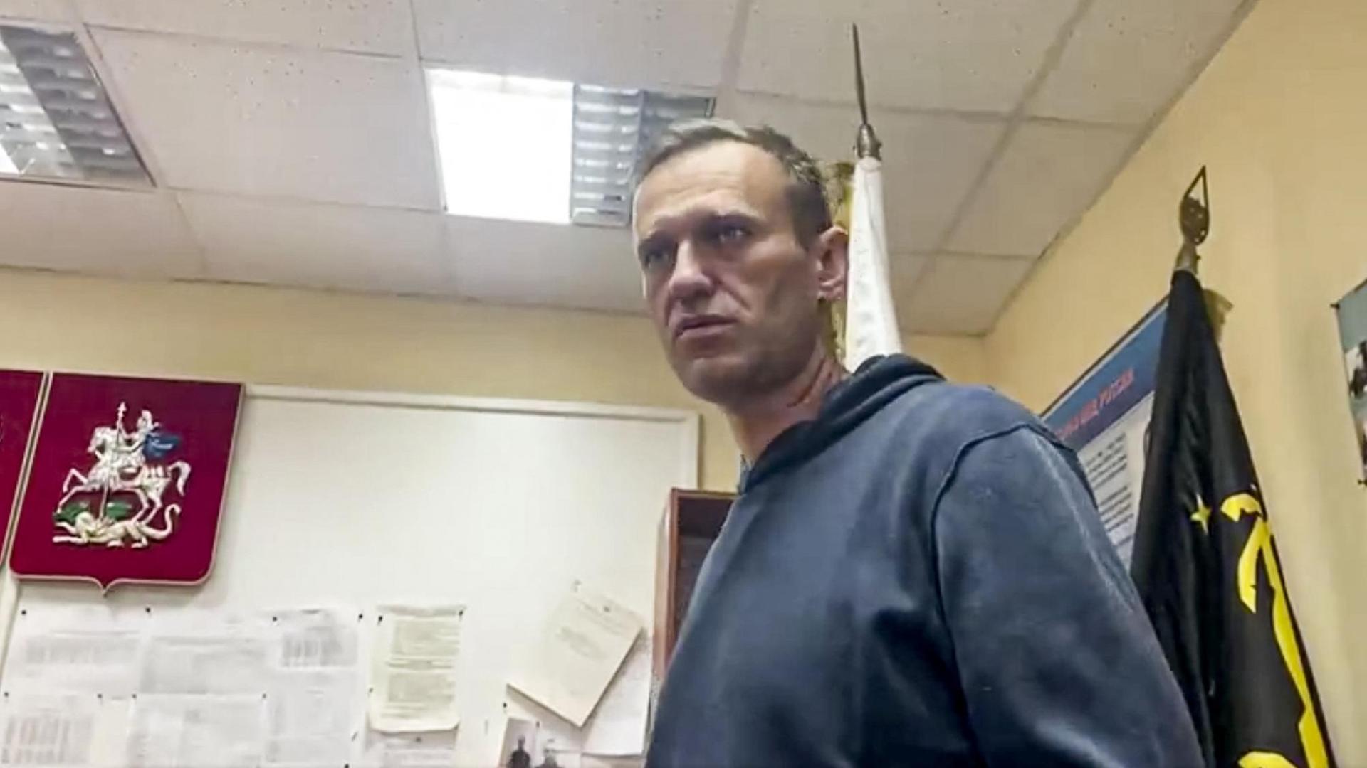 Nawalny im dunklblauen Pulli ist von unten fotografiert. Er steht in einem Dienstzimmer mit Fahnen und Wappen an der Wand und blickt ernst. Nawalny im dunklblauen Pulli ist von unten fotografiert. Er steht in einem Dienstzimmer mit Fahnen und Wappen an der Wand und blickt ernst.