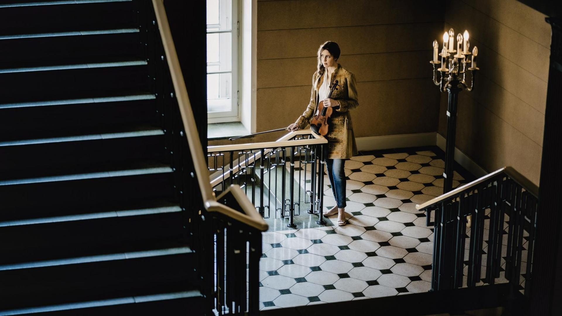 Eine junge Frau steht mit Geige und Bogen in einem Treppenhaus.