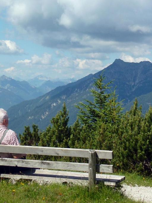 Ein älterer Herr sitzt auf einer Bank vor einem Bergpanorama