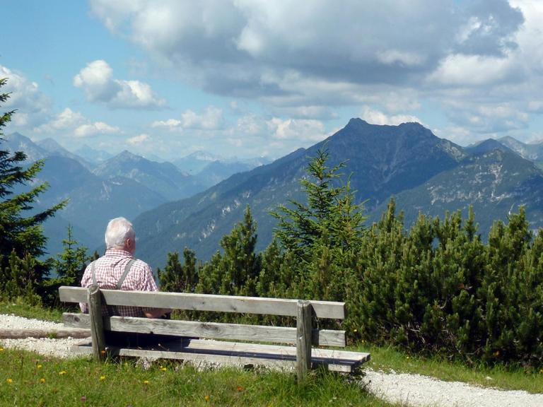 Ein älterer Herr sitzt auf einer Bank vor einem Bergpanorama