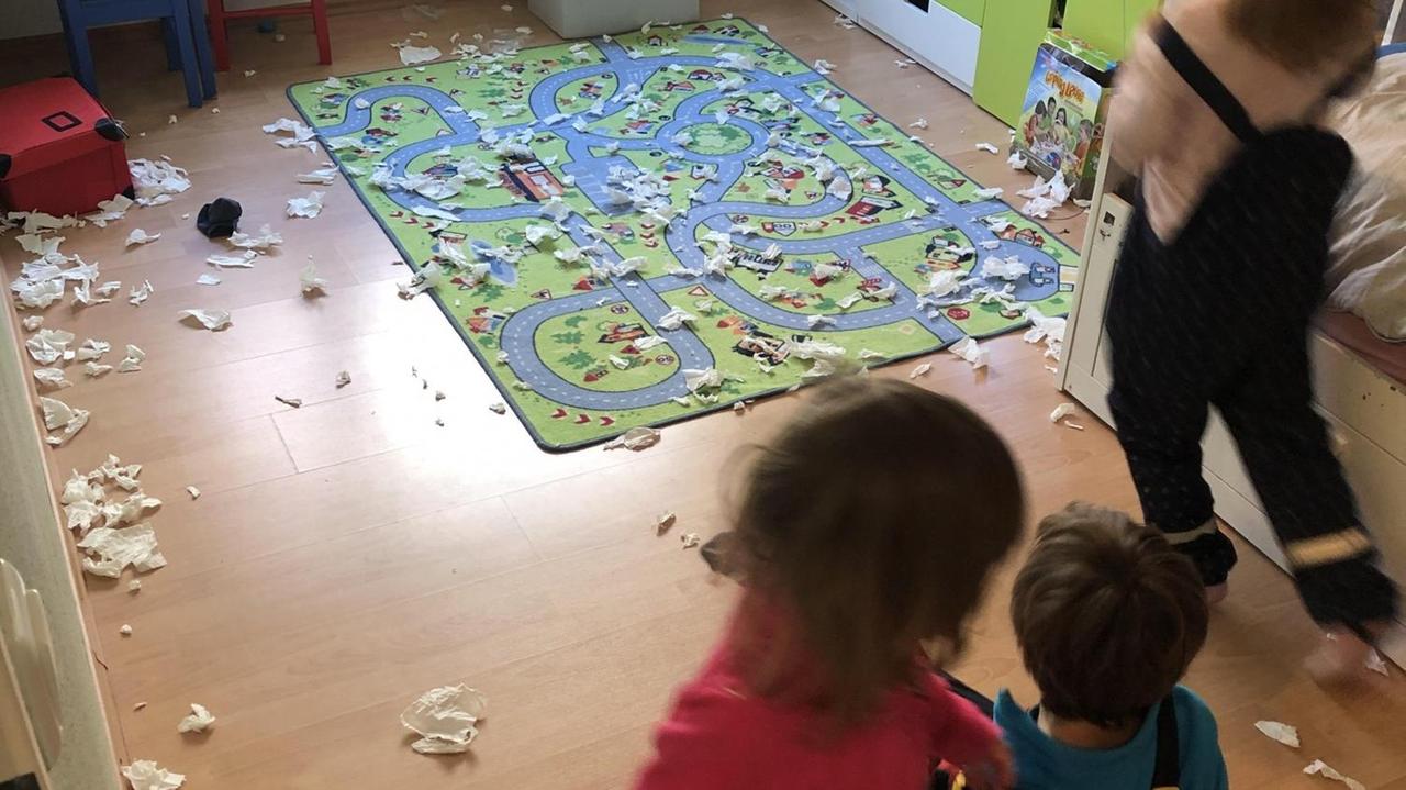 Der Fußboden des Kinderzimmers ist mit einem Spielteppich und vielen Papierschnipseln bedeckt. Zu sehen sind unscharf drei kleine Kinder. Der Fußboden des Kinderzimmers ist mit einem Spielteppich und vielen Papierschnipseln bedeckt. Zu sehen sind unscharf drei kleine Kinder.