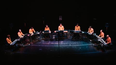 12 Musiker in orangefarbenen Hemden sitzen im Halbkreis auf der Bühne